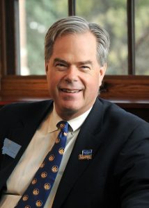A smiling man with gray hair in a white-button up shirt, blue tie with gold, circular accents, and black jacket with pin and Montana emblems on his lapels